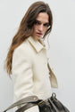 A person with long brown hair wearing a veste élégante cream-colored coat looks over their shoulder while holding a dark gray bag.