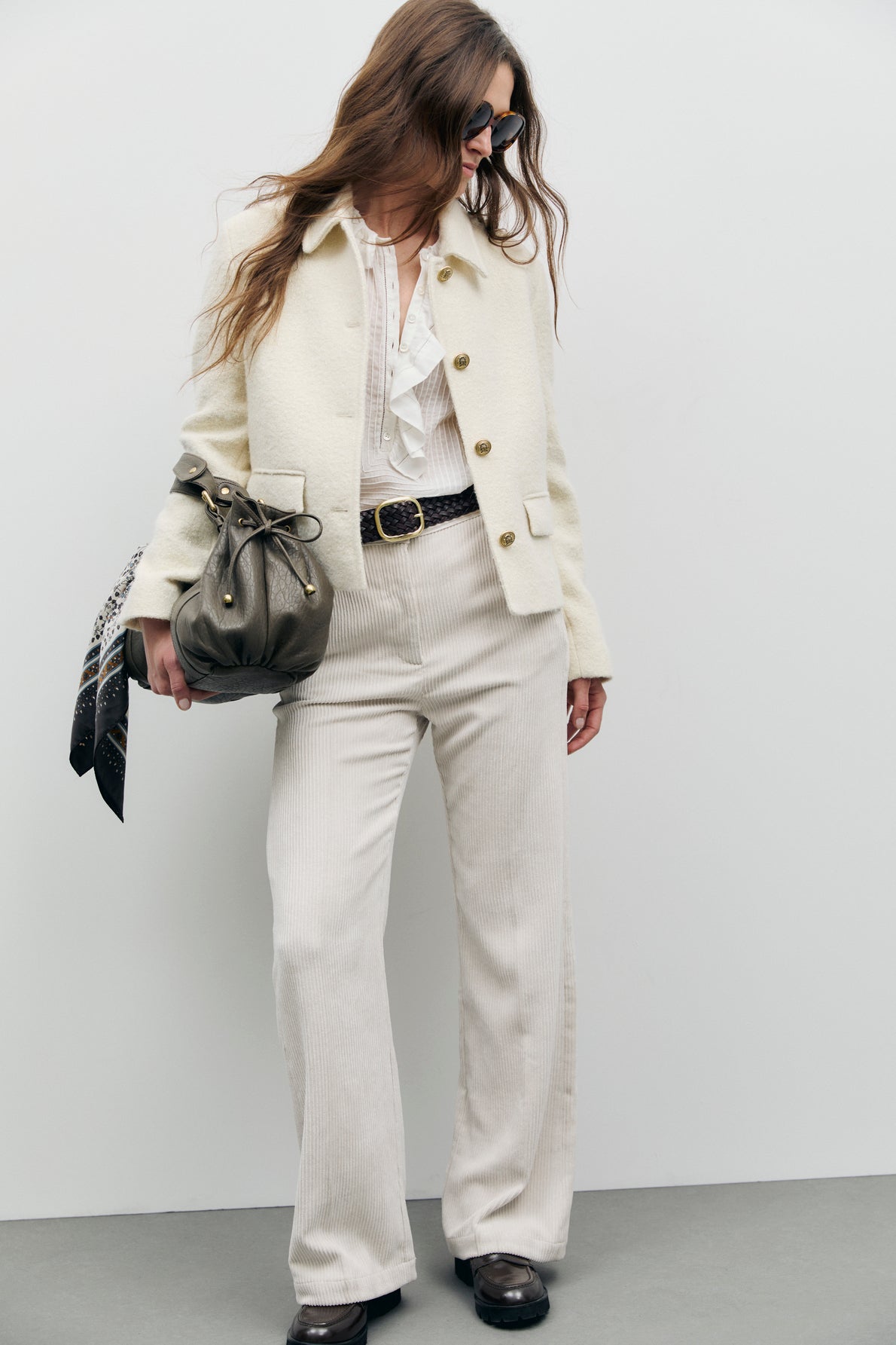 A woman in sunglasses wears a veste élégante écru, a white ruffled blouse, high-waisted cream pants, and dark shoes. She carries an olive bag with a tied scarf and has long, wavy hair.
