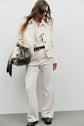 A woman in sunglasses wears a veste élégante écru, a white ruffled blouse, high-waisted cream pants, and dark shoes. She carries an olive bag with a tied scarf and has long, wavy hair.