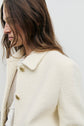 A woman with long brown hair wears a veste courte écru, a cream-colored textured coat with gold buttons, over a light blouse, standing against a plain light background. Her face is partially turned away.
