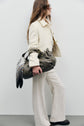A woman in cream-colored pants and a matching veste élégante walks, carrying a large olive green bag with a patterned scarf. She has long brown hair and wears sunglasses.
