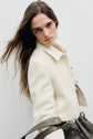 A woman with long brown hair wears a veste courte écru and holds a dark brown bag. She stands against a plain white background, looking directly at the camera with a neutral expression.