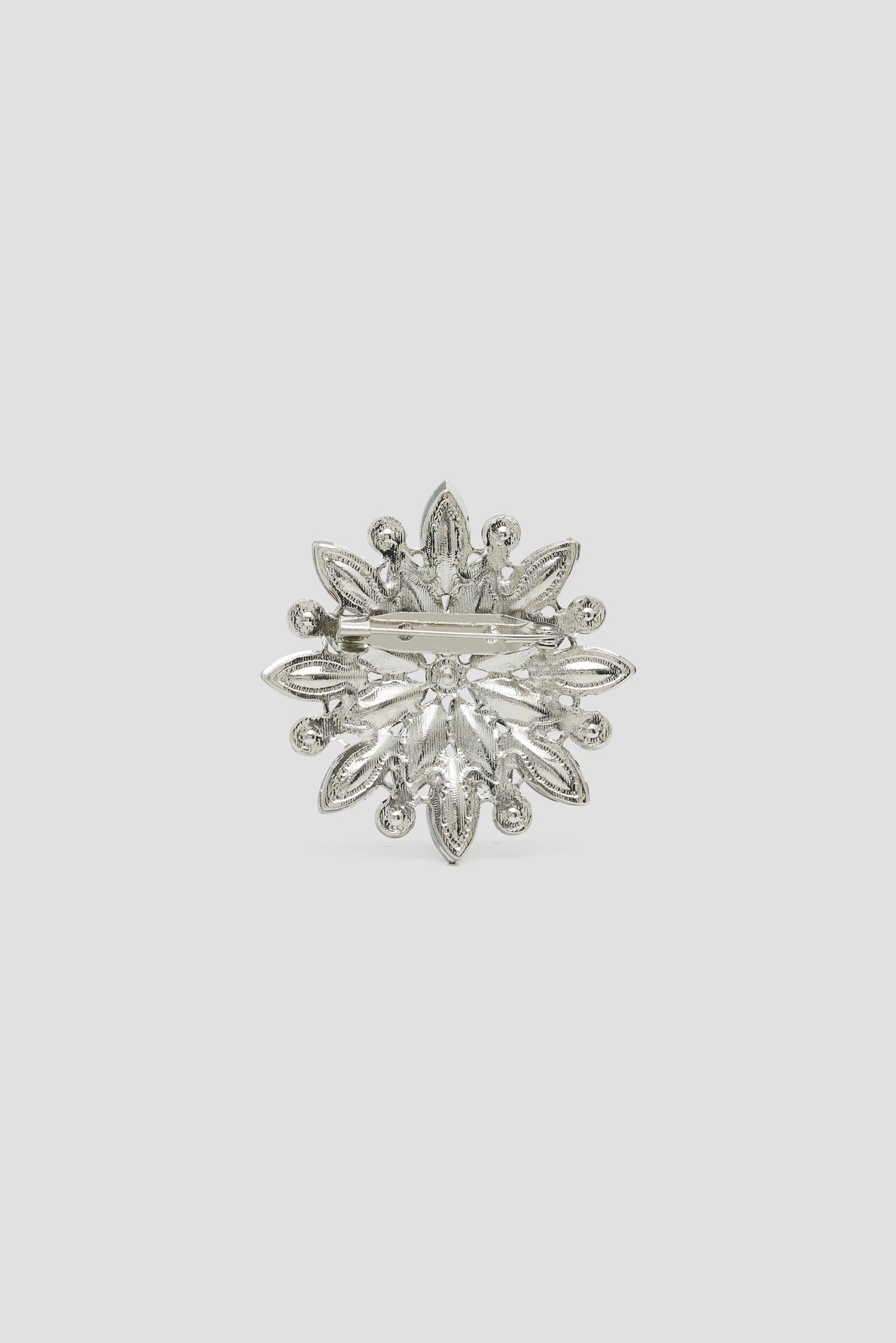 Silver-toned brooch with a floral, snowflake-like design, featuring petal-shaped and round decorative elements, shown against a plain light gray background. This broche élégante has a pin clasp visible on the back, making it an ideal accessory for adding refined charm to your veste.