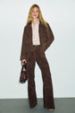 A person with long blonde hair wears a brown suede jacket and matching pantalon marron femme flare cuir velours, paired with a beige shirt, and holds a brown handbag, standing against a light background.