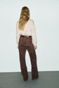 A person with long, wavy hair is standing with their back to the camera, wearing a beige long-sleeve shirt paired with a black belt and a pantalon flare cuir velours marron. The vintage-inspired look is set against a plain light background, highlighting the timeless elegance of this pantalon marron femme.