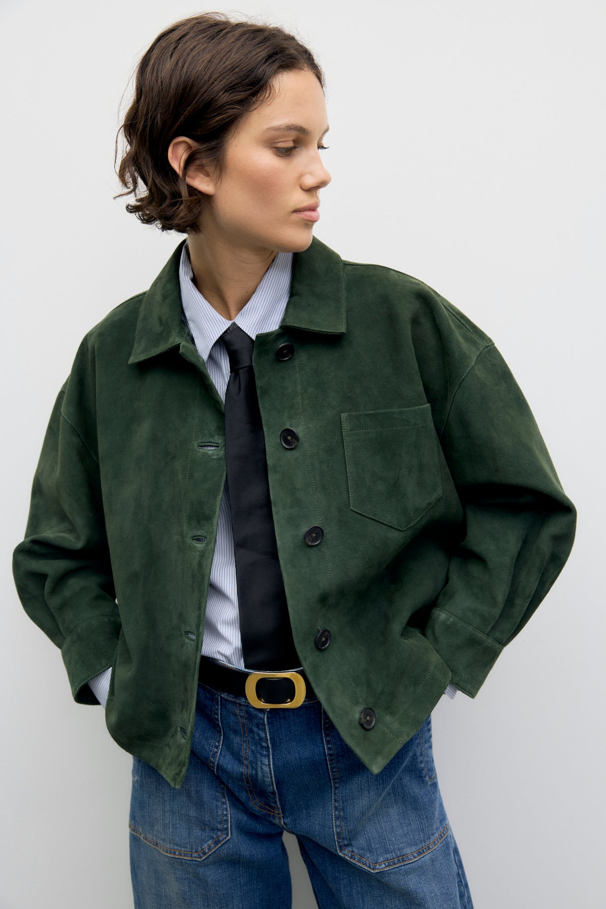 A person wearing a green veste courte en cuir velours, striped shirt, black tie, blue jeans with a belt, stands with hands in pockets, looking to the side against a plain light background.