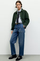 Person standing against a white wall, wearing a green veste courte over a light blue shirt, black tie, high-waisted blue jeans with a black belt, and black loafers. They have short brown hair and a neutral expression.