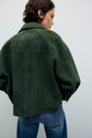 A person with short brown hair wears a loose, dark green cuir velours surchemise femme and blue jeans, facing away against a plain light background.