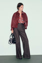 A person wearing a veste bordeaux in leather, paired with a floral blouse, wide-leg brown trousers, and black shoes, holding a black handbag, stands against a plain light green background.