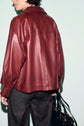 A person wearing a loose-fitting surchemise cuir bordeaux and dark pants, holding a black handbag, is shown from the back against a pale background.