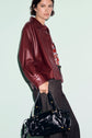 A person wearing a maroon leather jacket, styled as a surchemise cuir, and dark pants holds a shiny black handbag with gold hardware against a light background.