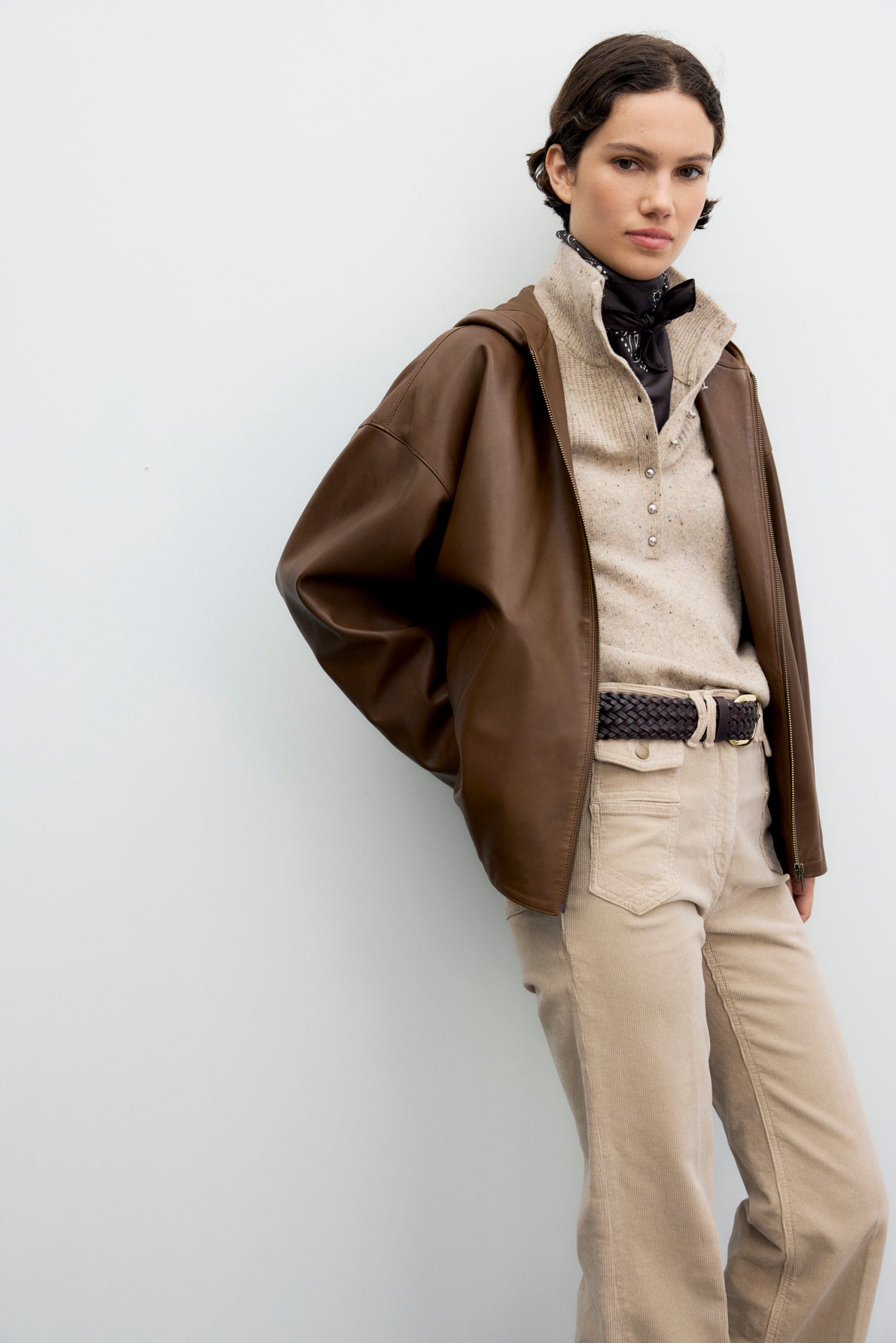 A person with short dark hair wears a brown veste en cuir, beige shirt, light pants, black scarf, and braided belt, standing against a plain light background.