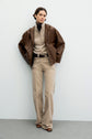 Woman stands against a light wall wearing a brown veste en cuir, beige buttoned sweater over a black top, beige wide-leg pants, and brown shoes. Hands are in pockets and she has short, wavy hair.