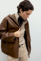 Person with short dark hair wearing a veste en cuir marron over a beige sweater and beige pants, looking down with one hand pulling the jacket open. They have a dark scarf around their neck.
