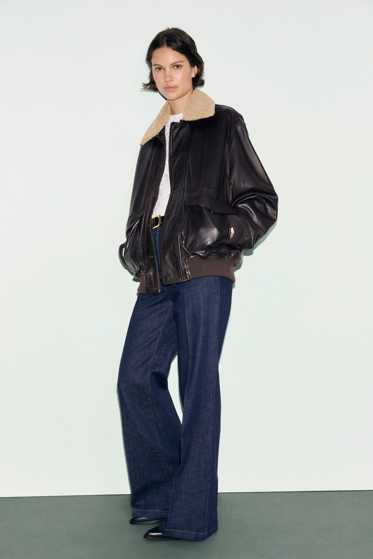 A person stands against a plain background, wearing a dark brown blouson en cuir with a tan col mouton shearling collar, blue wide-leg jeans, and black shoes.