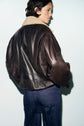 Person with short dark hair wearing a brown blouson en cuir with a col mouton and blue jeans, standing against a light background, facing away from the camera.
