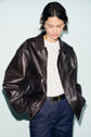 A person wearing a dark brown blouson aviateur en cuir featuring a col mouton over a white patterned top and blue jeans with a black belt, looking down with hands in jacket pockets against a light background.