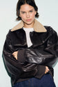 A person wearing a dark blouson aviateur en cuir with a beige col mouton and a white knit top stands against a light background, looking confidently at the camera.