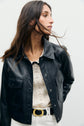 Woman with long brown hair wearing a veste cuir noir courte over a white lace top and white pants with a black and gold belt, looking to the side against a plain light background for an effortlessly chic style vintage look.