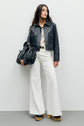A woman stands against a plain wall wearing a veste courte en cuir noir in true style vintage, paired with white wide-leg pants, platform shoes, and holding a large black bag.