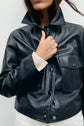 A person wearing a veste courte en cuir noir with large front pockets and a cream sweater sleeve visible, holding the jacket collar. The background is plain and light-colored, giving the look a subtle style vintage appeal.