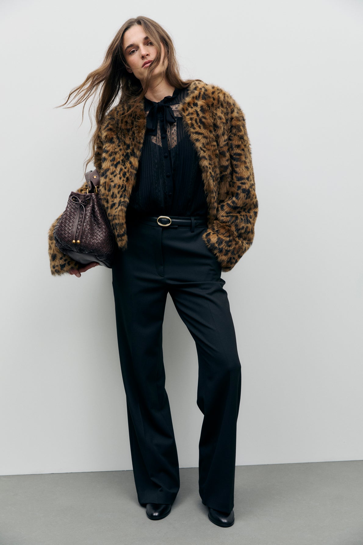 Woman stands against a white wall wearing a manteau court à imprimé léopard, black blouse, black wide-leg pants, and black shoes, holding a textured brown handbag.