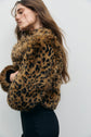 Woman with long brown hair wearing a manteau court imprimé léopard faux fur jacket and black pants, standing sideways against a plain light-colored background.