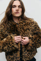 A person with long brown hair wears a manteau court imprimé léopard en fausse fourrure, looking directly at the camera against a plain light background.