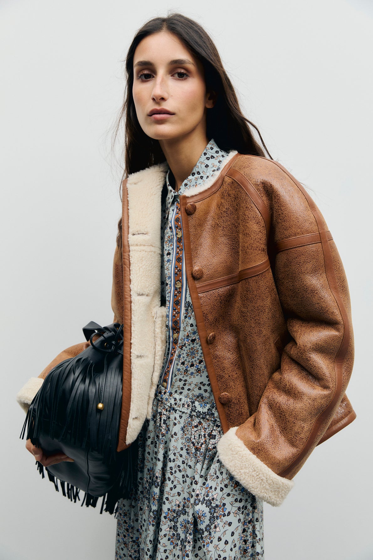 A woman with long dark hair wears a blouson cuir agneau over a floral dress and holds a black fringe handbag, standing against a plain light background.