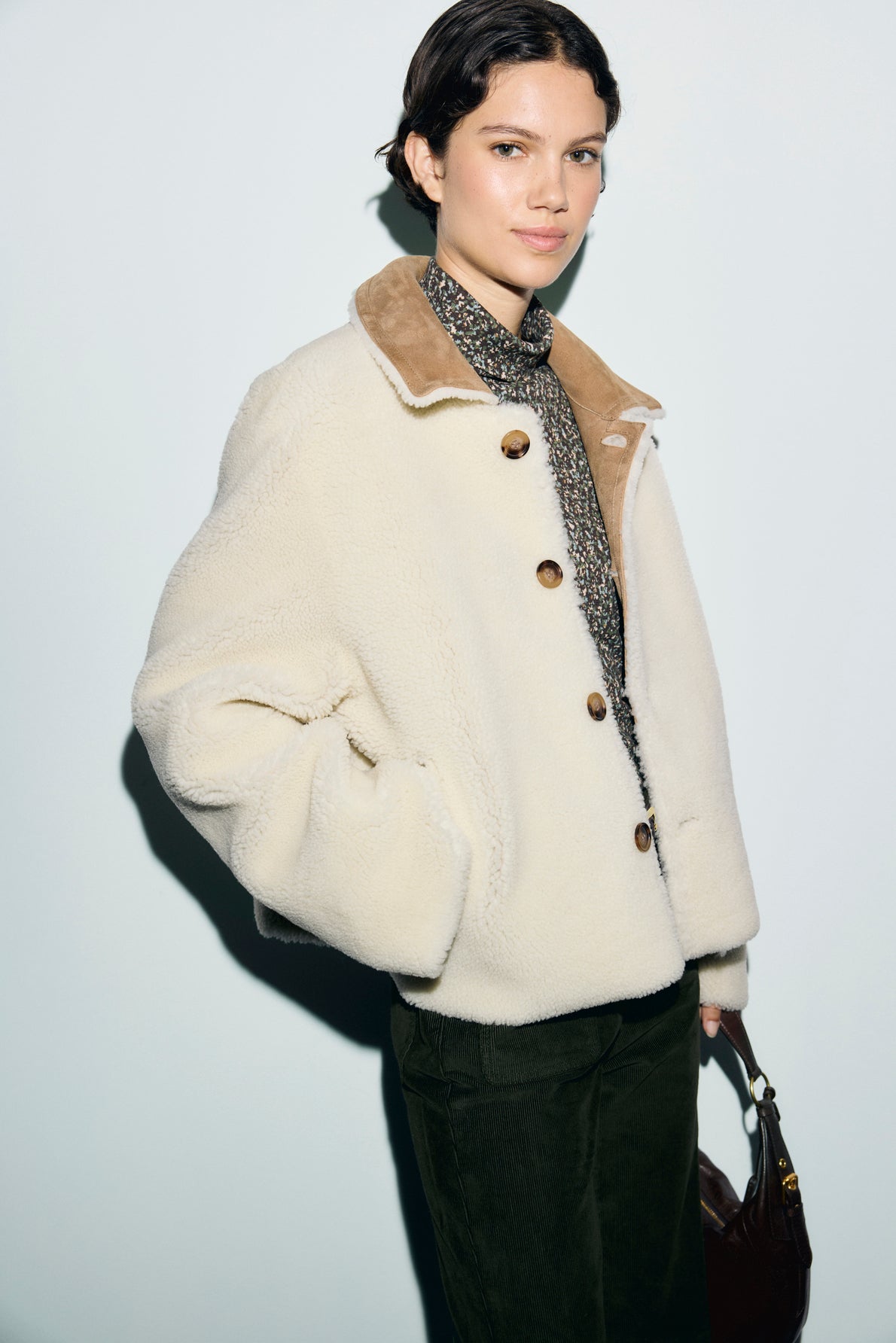 A person wearing a cream-colored peau lainée shearling jacket with brown buttons, a patterned scarf, and dark pants stands against a light background, holding a bag.