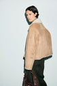 A person with short dark hair wears a tan peau lainée shearling jacket and dark pants, holding a brown bag, standing against a light background and looking slightly over their shoulder.
