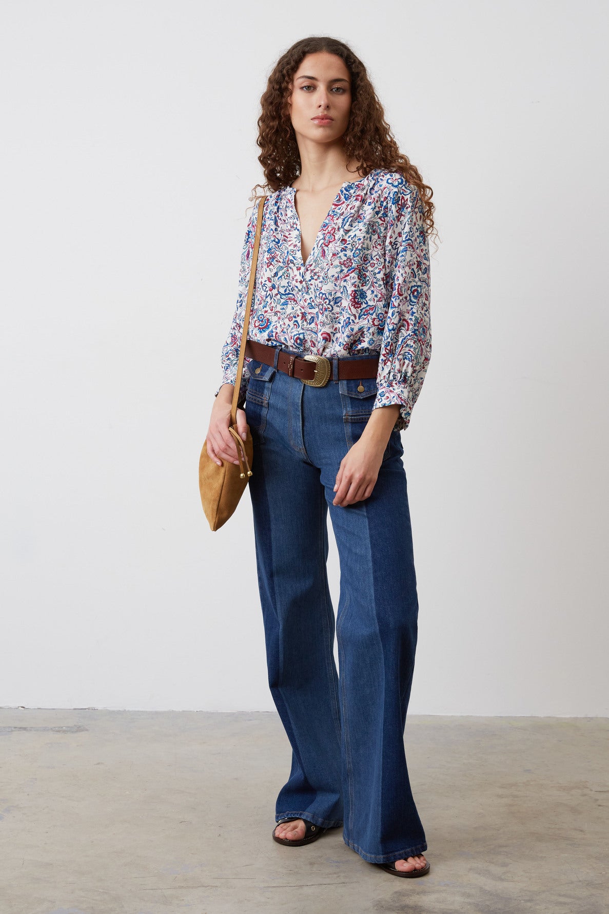 A woman with long curly hair wears a patterned blouse, high-waisted flared jeans, a wide belt, black sandals, and carries a tan shoulder bag, standing against a plain white wall on a concrete floor.