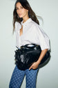 Woman with long brown hair wearing a white chemise femme in popeline de coton and blue patterned jeans, holding a large black leather bag under her arm, standing against a light background.
