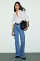 Woman stands against a plain background, wearing a chemise oversize blanche in crisp popeline de coton, paired with patterned blue flared jeans, black shoes, and holding a large black handbag on her shoulder.