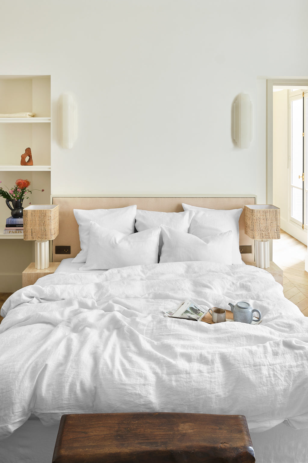 A neatly made bed with lin lavé blanc linens, pillows, and a tray holding a teapot, cup, and book. Two lamps with woven shades are on side tables, and shelves with flowers, books, and decor are in the background. The room’s serene atmosphere is enhanced by the luxurious housse de couette certifiée for a touch of effortless elegance.