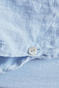 Close-up of a light blue linen shirt, showing a white button partially fastened and detailed fabric texture.