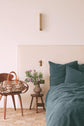 A cozy bedroom with a teal bedding set, a small wooden stool with a vase of flowers, a wall sconce, and a wooden chair holding a patterned handbag. The decor is minimal and modern.