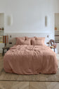 A neatly made bed with blush pink bedding and pillows stands in a minimalist bedroom with neutral walls, two side tables, soft lighting, shelves, and a woven rug on light wooden floors.