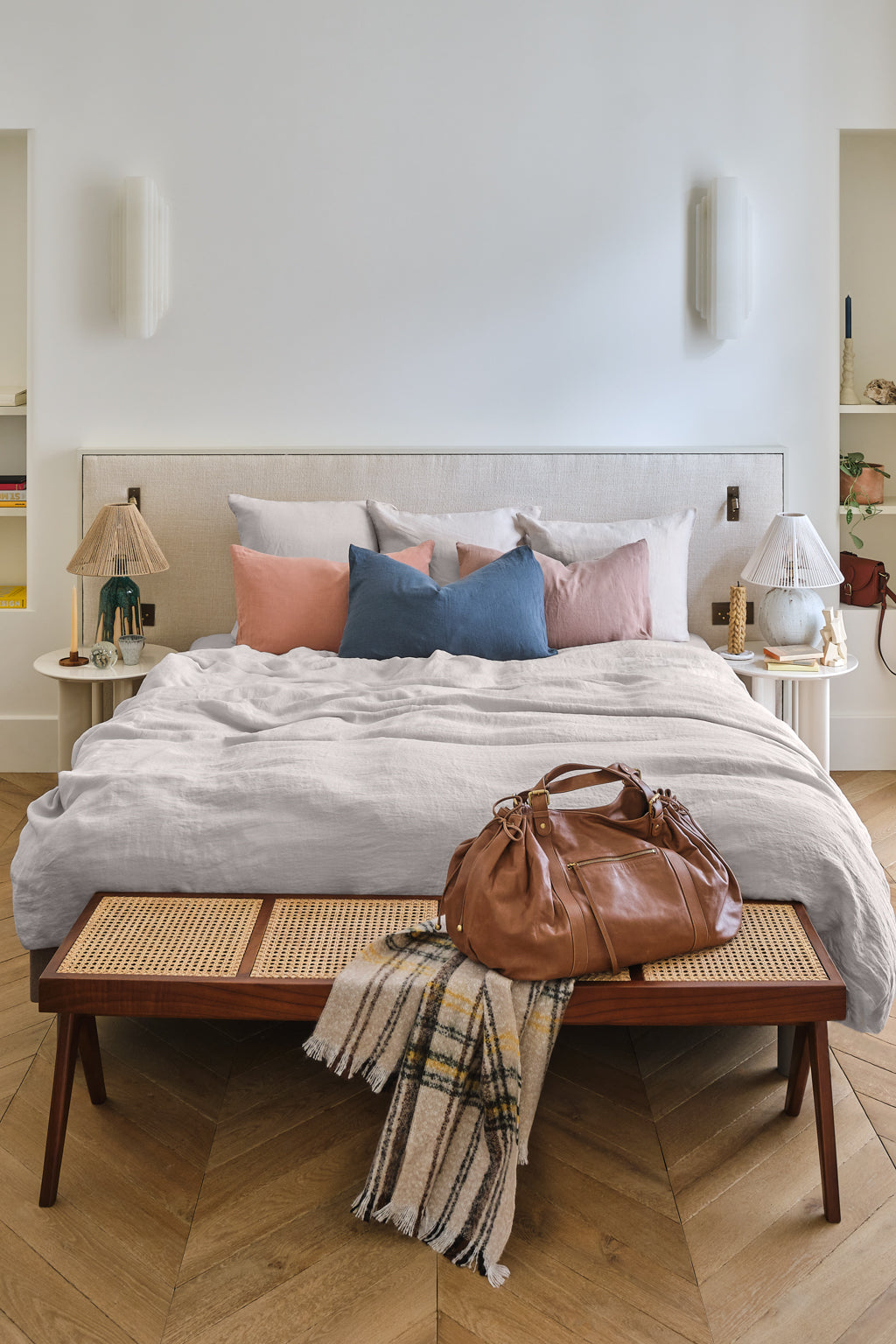 A cozy bedroom with a bed adorned with pink, blue, and gray pillows, flanked by two side tables and lamps. A brown leather bag and a plaid blanket rest on a wooden bench at the foot of the bed.