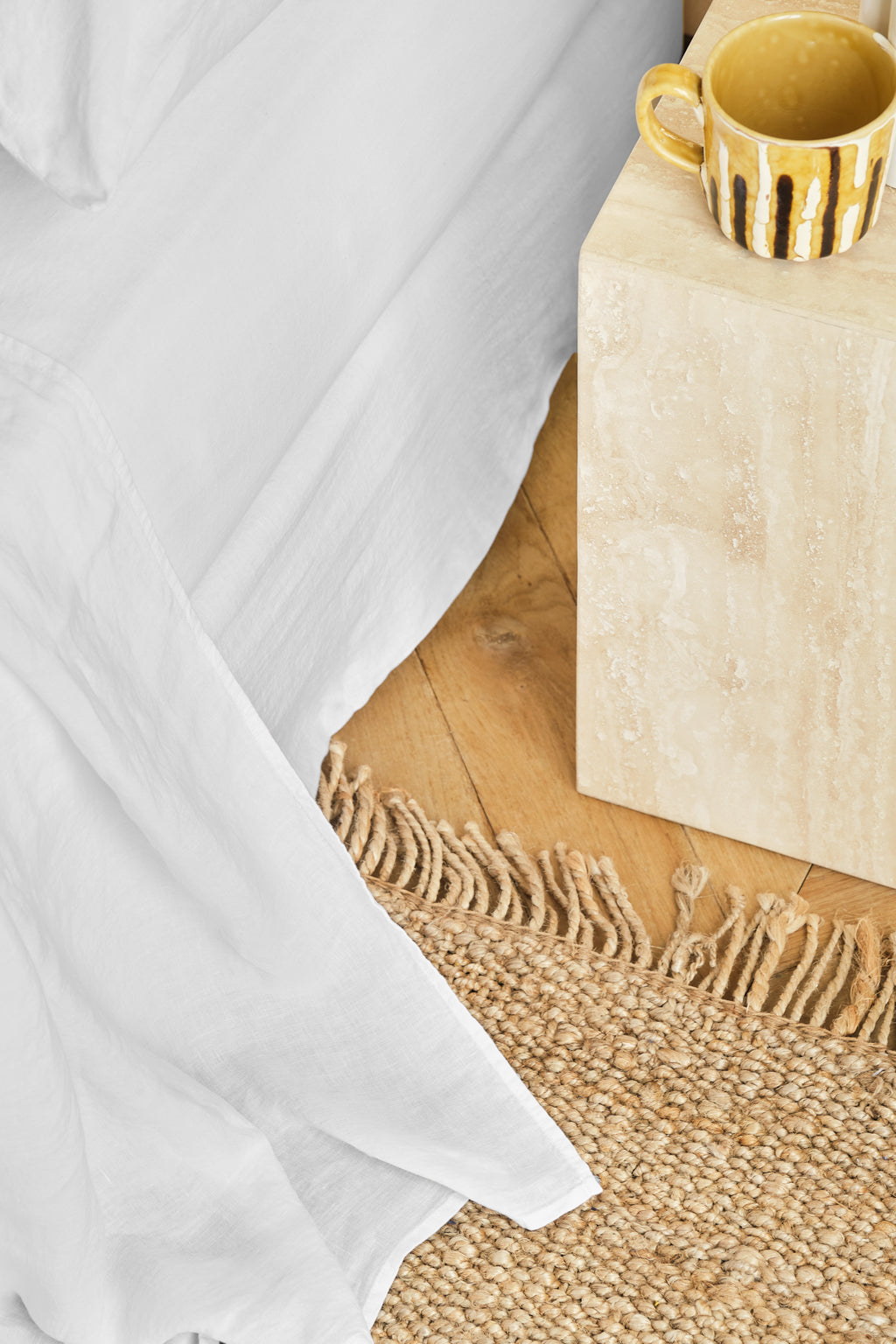 A yellow mug with brown stripes sits on a beige stone bedside table next to a white bedspread and a woven straw rug on a wooden floor.