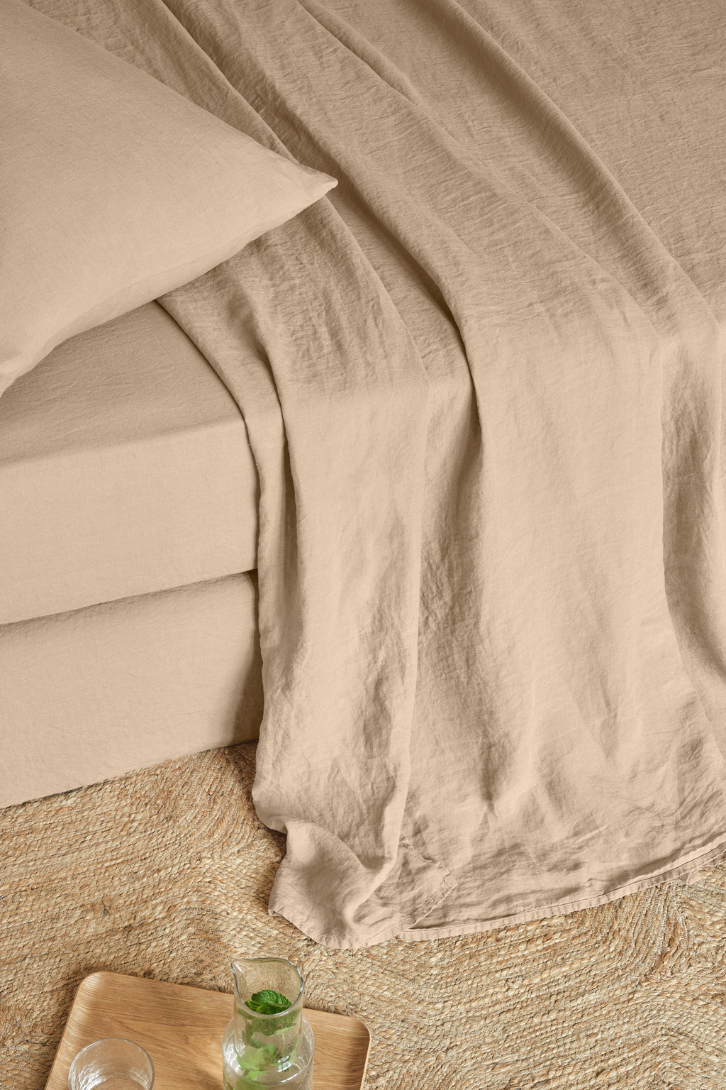Beige bedding with a pillow and sheet neatly arranged on a bed, placed on a woven rug. A wooden tray with a glass of water and a small bowl sits on the floor nearby.