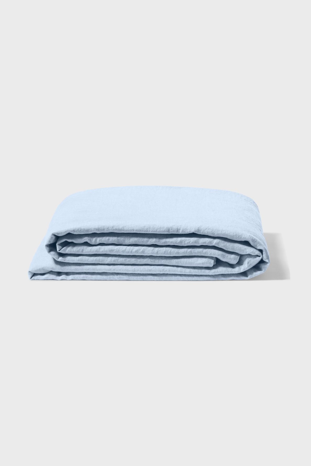 A drap plat lin lavé bleu ciel, certifié OEKO-TEX, est soigneusement plié et empilé sur un fond gris clair et épuré.
