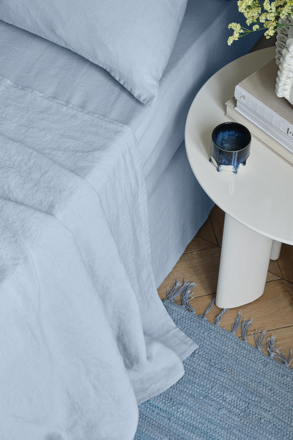 A neatly made bed with bleu ciel linen, including a drap plat lin lavé certifié OEKO-TEX, sits next to a white round side table holding a blue cup, books, and a vase of yellow flowers. A textured blue rug is on the wooden floor beside the bed.