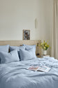 A neatly made bed with light blue bedding in a soothing bleu ciel shade, featuring a drap plat lin lavé certifié OEKO-TEX, four pillows, an open magazine on the duvet, a vase of yellow flowers on the nightstand, and a framed animal photo on the headboard against a cream wall.
