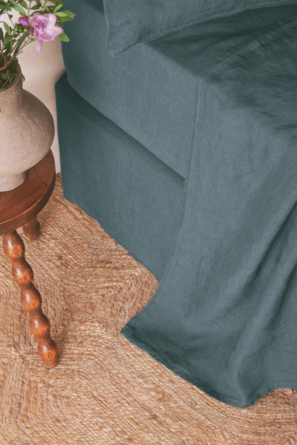 A corner of a teal bed with linen bedding next to a wooden side table holding a vase with purple flowers, on a woven jute rug.