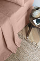 A bed with blush pink linen sheets and pillow, beside a wooden table holding a ceramic jug and books. A woven rug with fringed edges lies on the wooden floor.