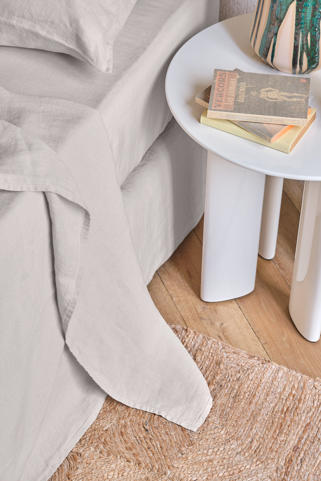 A neatly made bed with light gray bedding is next to a round white side table holding books and a lamp. A woven rug is on the wooden floor.