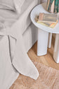 A neatly made bed with light gray bedding is next to a round white side table holding books and a lamp. A woven rug is on the wooden floor.