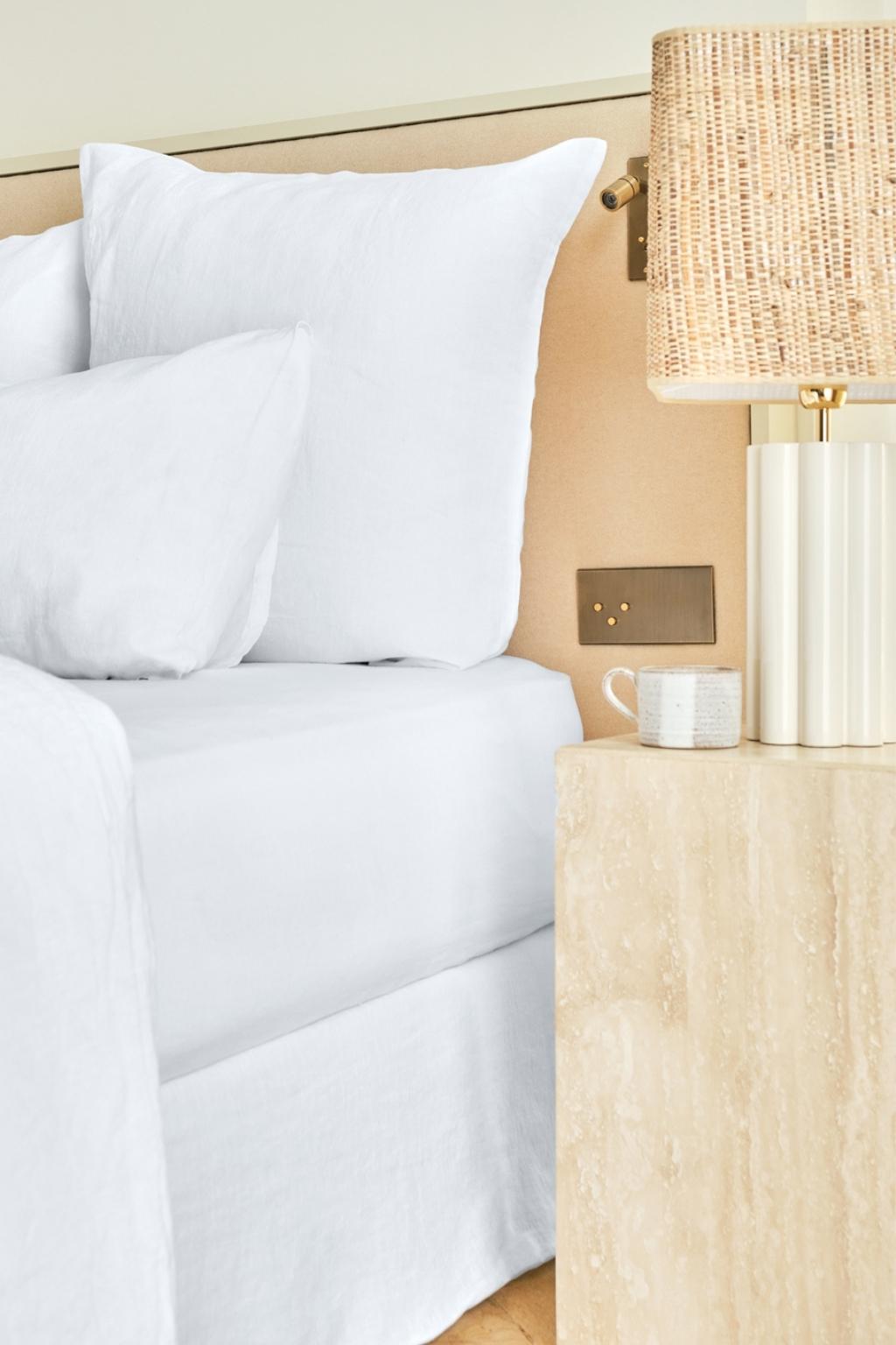 A neatly made bed with white linens next to a beige marble nightstand holding a textured lamp and a white mug. The scene is bright and minimalistic.