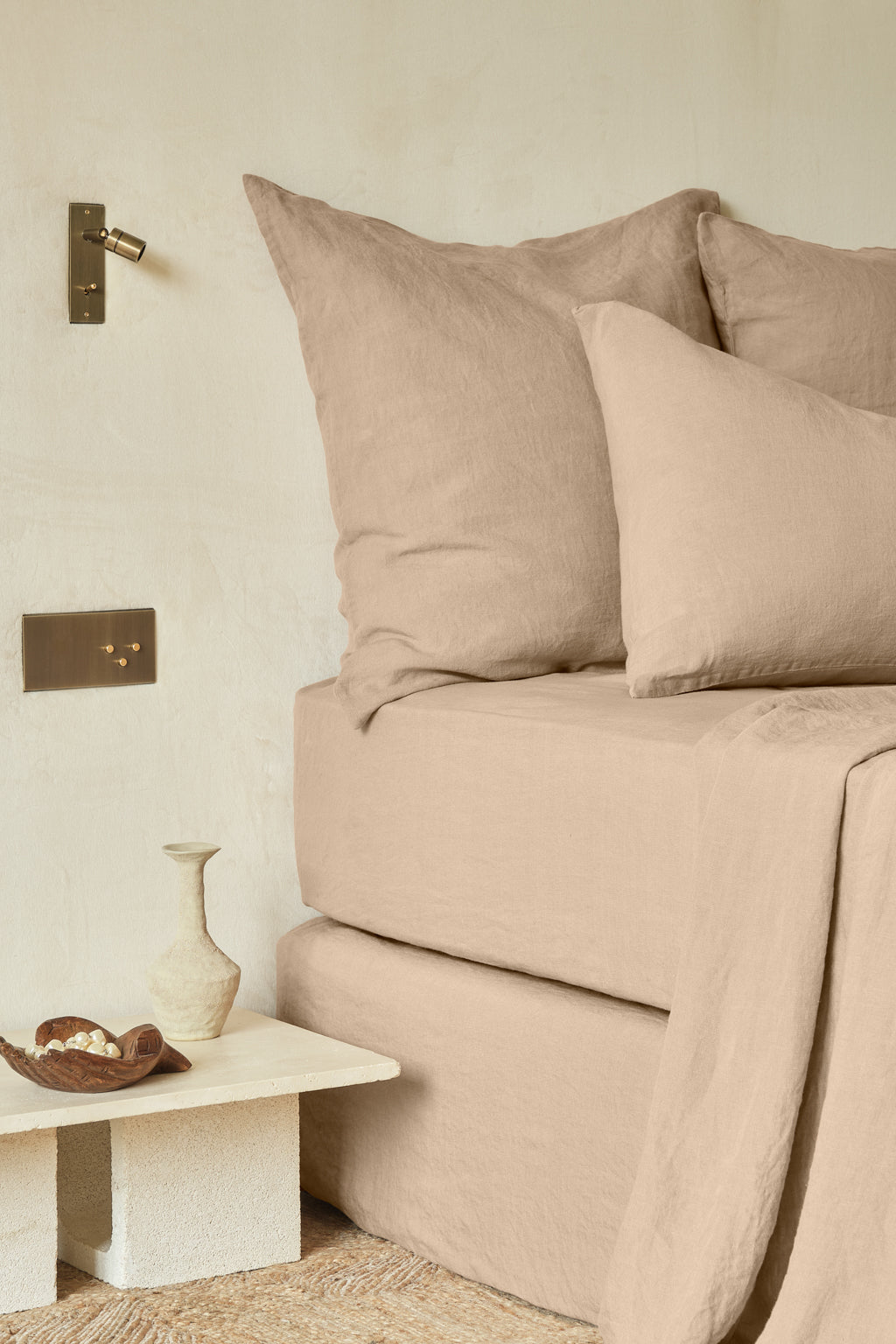 A neatly made bed with beige linen bedding, large pillows, and a matching sheet beside a small table holding a ceramic vase and a dish, set against a neutral wall with brass light fixtures.